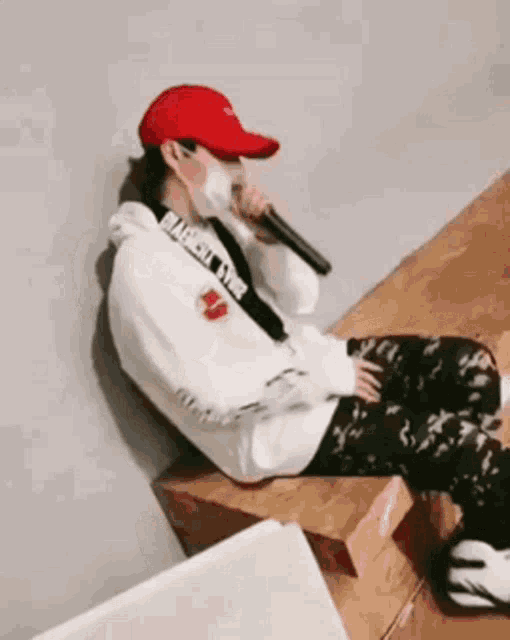 a person wearing a red hat and a mask is sitting on a wooden bench holding a microphone .