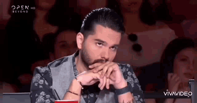 a man with a beard is sitting in front of a crowd with his hands on his face .