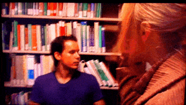 a man in a purple shirt is talking to a woman in front of a bookshelf