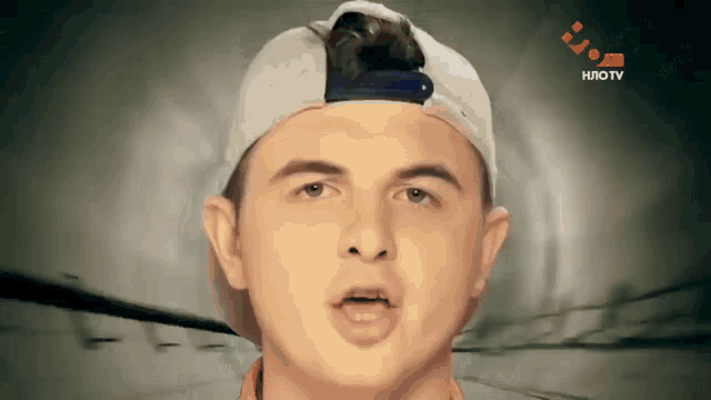 a young man wearing a baseball cap looks at the camera with a tv screen behind him that says hlotv