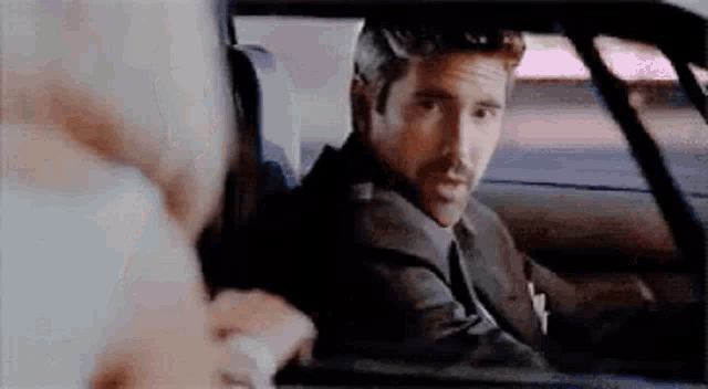 a man in a suit and tie is sitting in the back seat of a car talking to another man .
