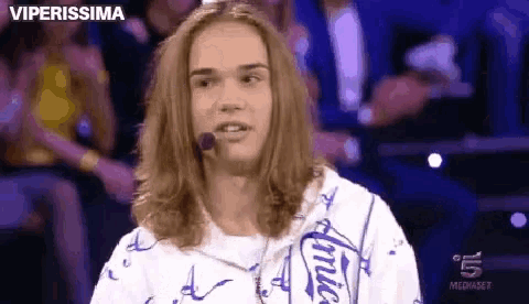 a young man with long hair is talking into a microphone while wearing a white shirt .
