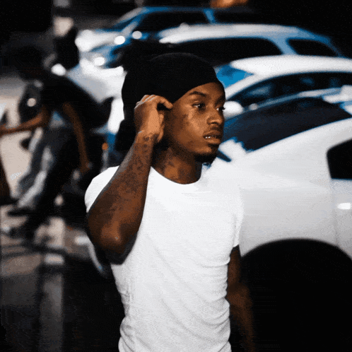 a man wearing a white shirt and a black beanie stands in front of a white car