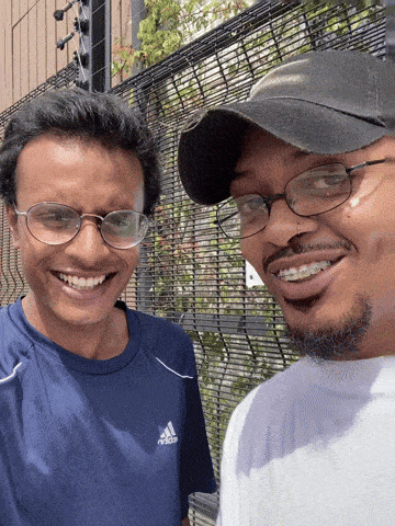 a man wearing a blue adidas shirt smiles next to another man wearing glasses