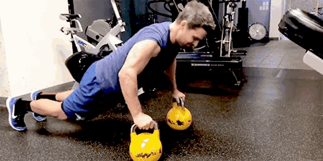 a man in a blue shirt is doing push ups with two yellow kettlebells in a gym