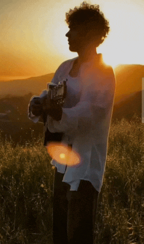 a man in a white shirt holds a guitar in a field at sunset