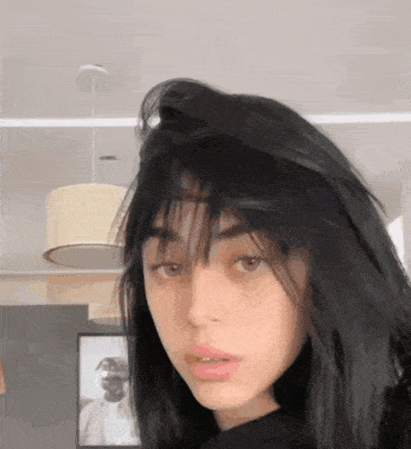 a girl with black hair looks at the camera with a picture of a man in the background