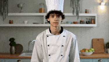 a young man in a chef 's hat stands in a kitchen looking at the camera