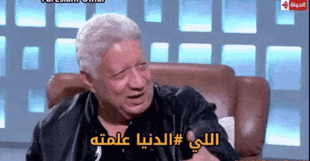 a man is sitting in a chair with arabic writing on the screen behind him