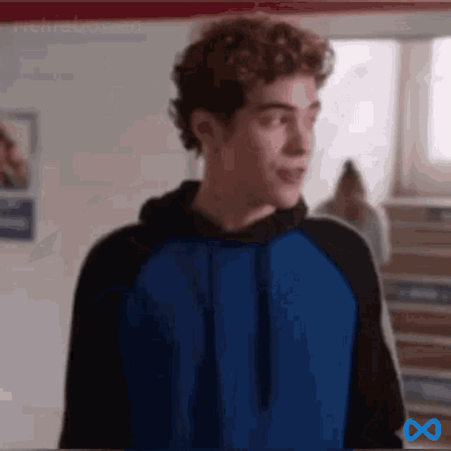 a young man with curly hair is wearing a blue and black hoodie and standing in a room .