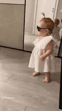 a baby girl wearing sunglasses and a white dress is standing on a wooden floor .