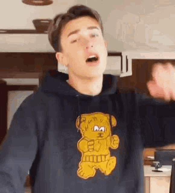 a young man wearing a black hoodie with a teddy bear on it is standing in a kitchen .