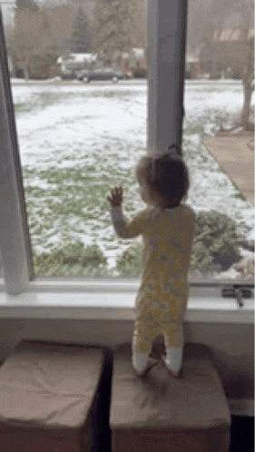 a baby stands on a window sill looking out at the snow