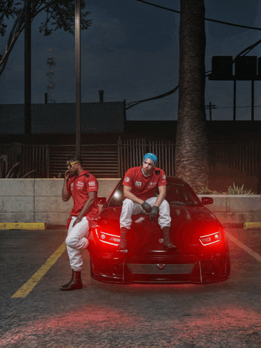 two men are sitting on a red car and one of them has a shirt that says ' a & t ' on it