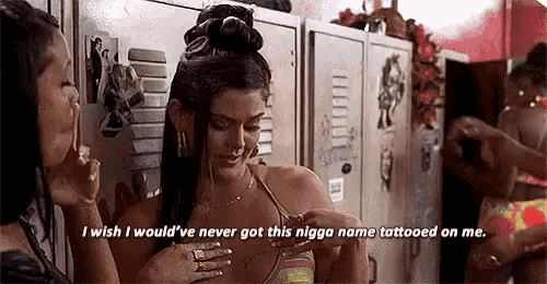two women are standing next to each other in a locker room . one of the women has a tattoo on her chest .