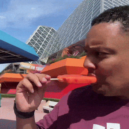 a man in a purple shirt is eating an orange popsicle on a stick