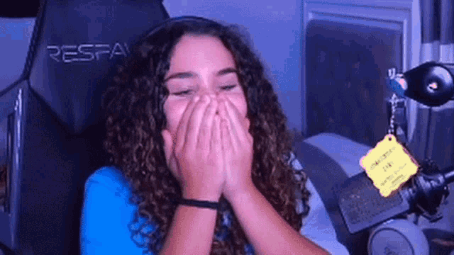 a woman with curly hair is covering her mouth with her hands while sitting in a chair in front of a microphone .