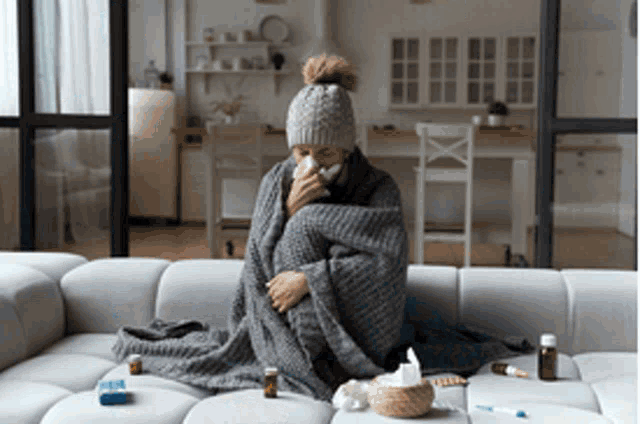 a woman wrapped in a blanket is sitting on a couch blowing her nose with a napkin