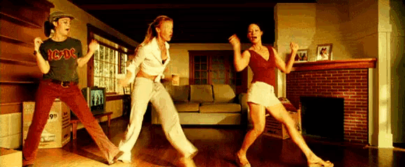 three women are dancing in a living room with a man in an ac dc shirt