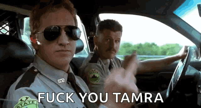 two police officers are driving a car and one of them is wearing sunglasses .