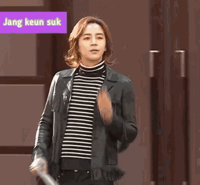 a young man wearing a striped sweater and a leather jacket is standing in front of a sign that says jang keur suk