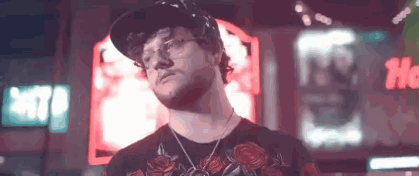 a man wearing glasses , a hat , and a necklace is standing in front of a neon sign .