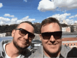 two men wearing sunglasses pose for a picture in front of a body of water