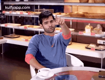 a man is sitting at a table in a bakery with a cup of coffee and making a funny face .