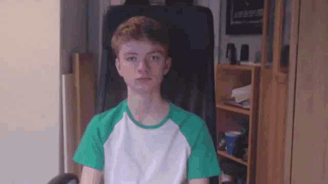 a young boy in a green and white shirt is sitting in an office chair looking at the camera .