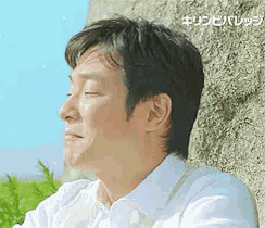 a man in a white shirt is leaning against a stone wall with his eyes closed .