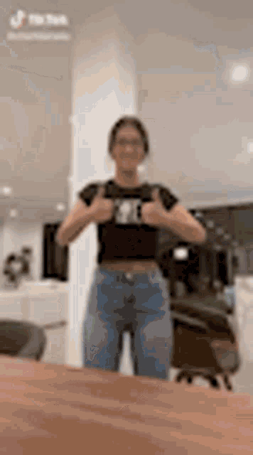 a woman in a black crop top and blue jeans is standing in front of a wooden table giving a thumbs up .