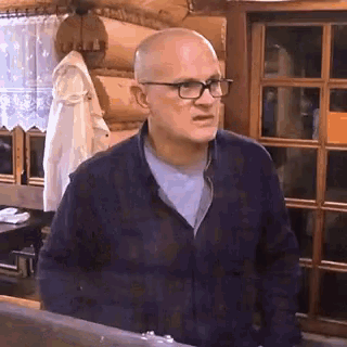 a bald man wearing glasses and a plaid shirt is standing in front of a window .