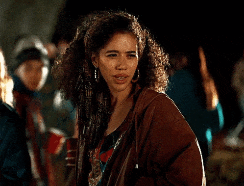 a woman with curly hair is wearing a brown jacket and earrings and standing in a crowd .