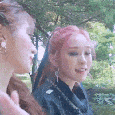 two women are standing next to each other in a park . one of the women has pink hair .
