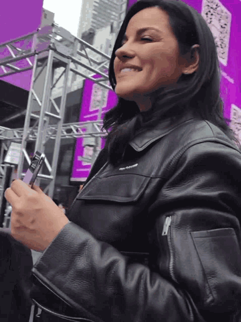 a woman in a black leather jacket is smiling while looking at her phone