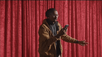 a man in a brown jacket is standing in front of a red curtain looking at his phone