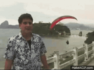 a man in a hawaiian shirt is standing in front of a body of water with a parachute in the background .