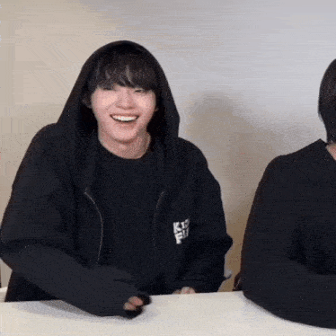 a man wearing a black hoodie is smiling while sitting at a table next to another man .