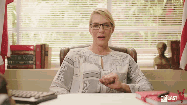 a woman wearing glasses sits at a desk with the word outcast on the bottom