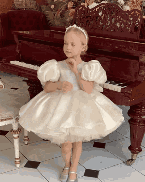 a little girl in a white dress stands next to a piano