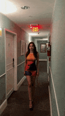 a woman walking down a hallway with an exit sign above her head