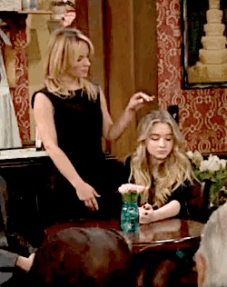 a woman in a black dress stands next to a girl sitting at a table with a vase of flowers