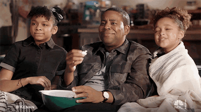 a man and two girls are sitting on a couch with a bowl of popcorn in front of them