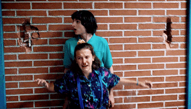mike and eleven are standing next to a brick wall .