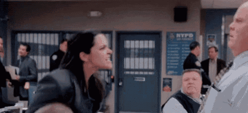 a woman is yelling at a man in a police station while a group of people are sitting around a table .