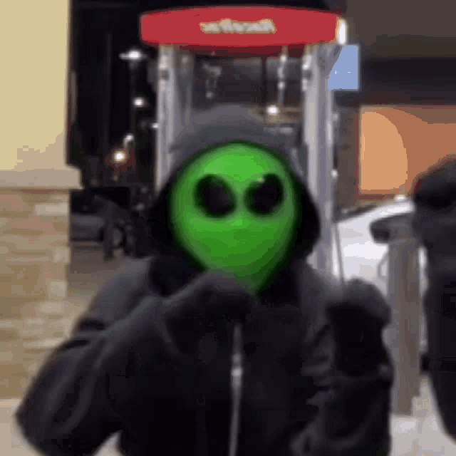 a person wearing a green alien mask and a hooded jacket is standing in front of a gas pump .
