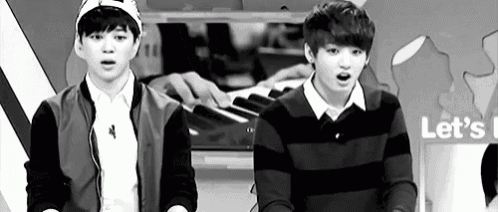 a black and white photo of two young men standing next to each other . one of the men is playing a piano .