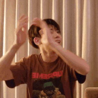 a man in a brown t-shirt is sitting on a couch with his arms outstretched in front of curtains .