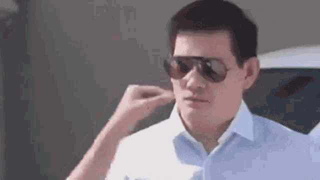 a man wearing sunglasses is touching his ear while standing next to a car .