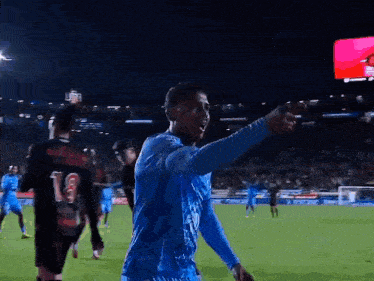 a man in a blue shirt is standing on a soccer field at night